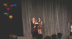 Two people standing on stage at Calgary Academy, one holding a microphone and the other receiving an award, with a dark curtain backdrop and audience in the foreground.