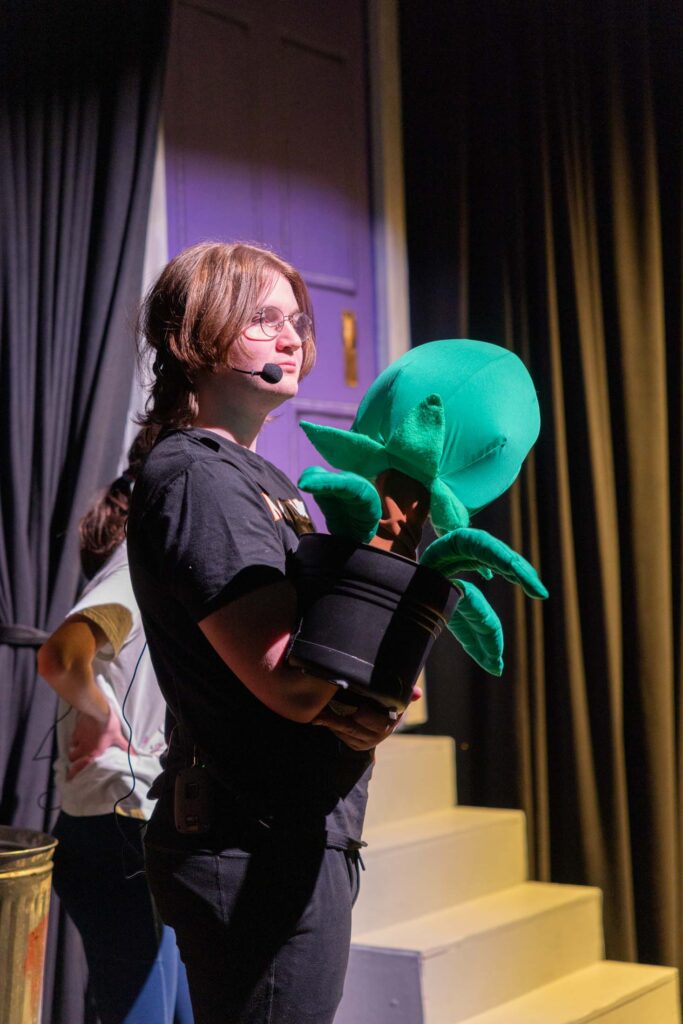 Calgary Academy student holding a green plant puppet on stage during the school play 'Little Shop of Horrors.'
