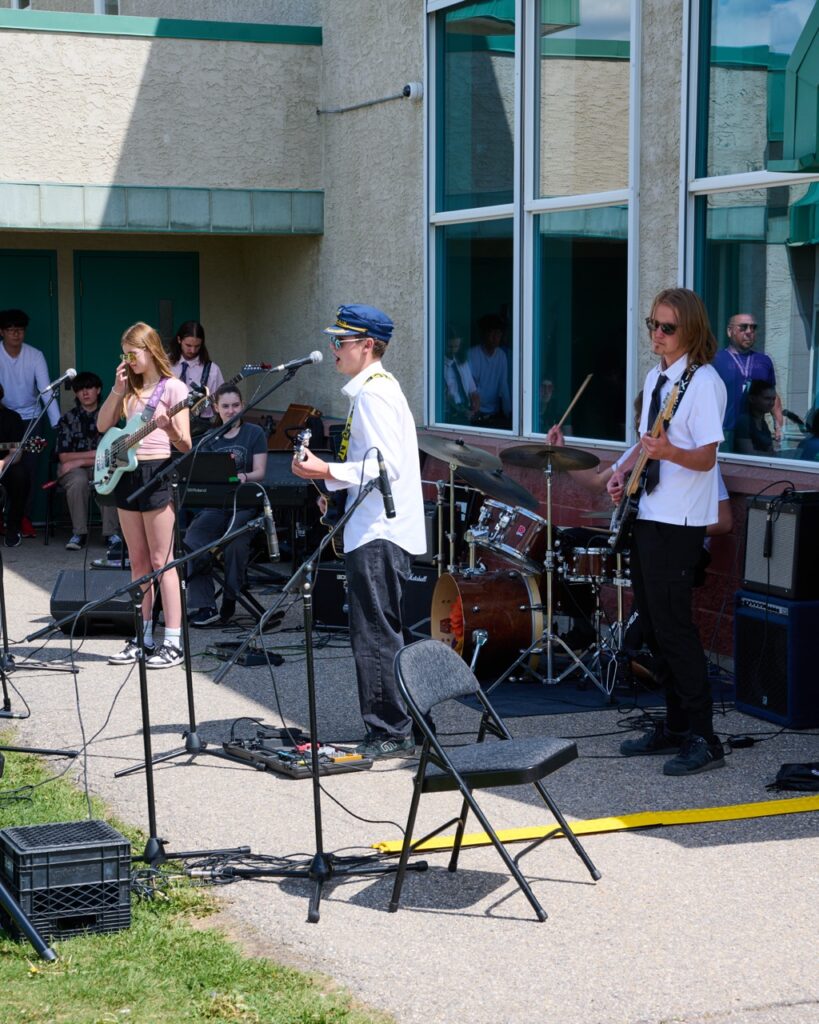 Calgary Academy student band performing outdoors at a school event, featuring electric guitar, bass, drums, and vocals, with an audience gathered in front of a school building.